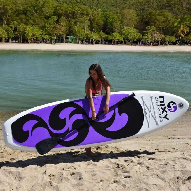 Women carrying a NIXY inflatable paddleboard and paddle in front of calm water and palm trees