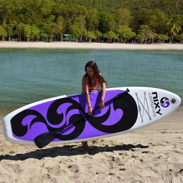 Women carrying a NIXY inflatable paddleboard and paddle in front of calm water and palm trees