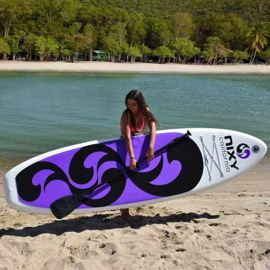 Women carrying a NIXY inflatable paddleboard and paddle in front of calm water and palm trees