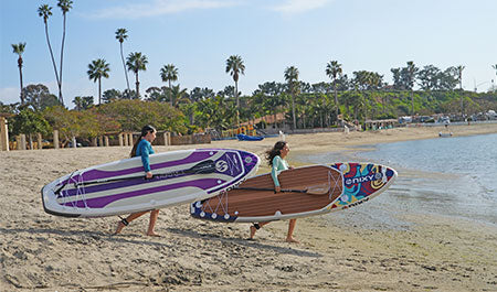 Two girls carrying inflatable paddleboards or iSUPs down to the water at the beach