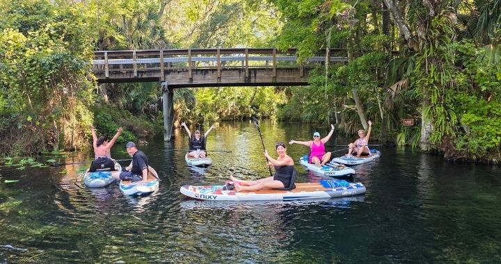 Family sitting on top of inflatable paddleboards in front of trees and a bridge