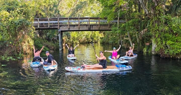Family sitting on top of inflatable paddleboards in front of trees and a bridge