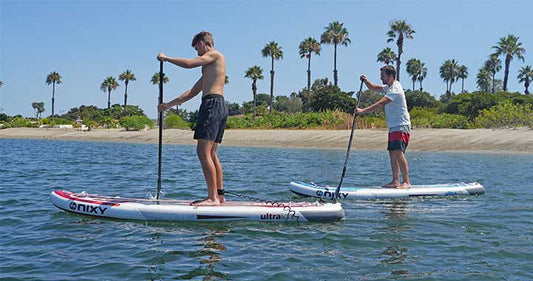 Men standing up on inflatable paddleboards in front of a scenic ocean view with palm trees and blue water.