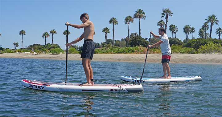 Men standing up on inflatable paddleboards in front of a scenic ocean view with palm trees and blue water.