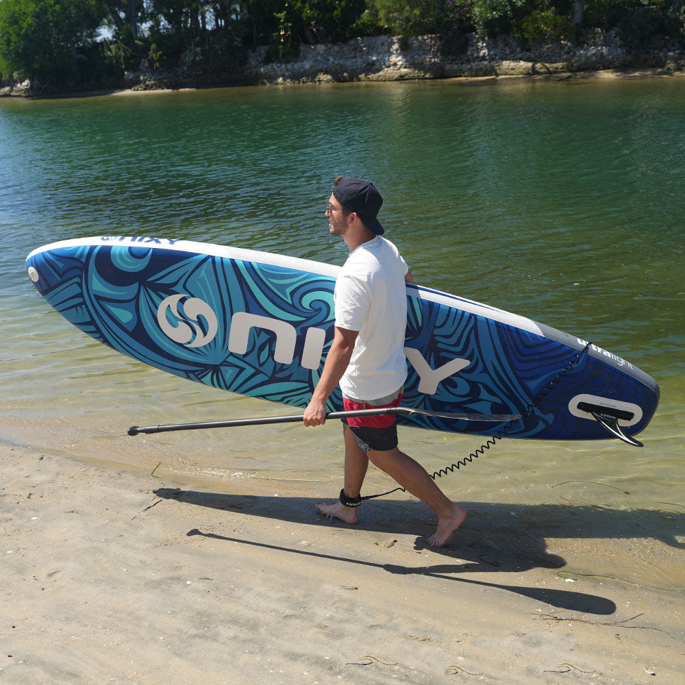 NIXY Newport G5 All-Around inflatable paddle board carried along a sandy shore with the included paddle