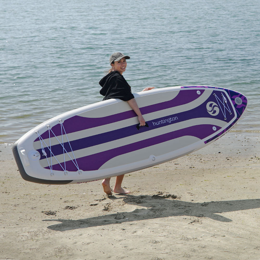 Woman carrying a NIXY Huntington G5 compact paddle board in purple across a sandy beach