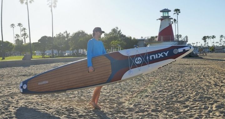 Man carrying inflatable paddleboard in front of a beach with palm trees and a lighthouse.