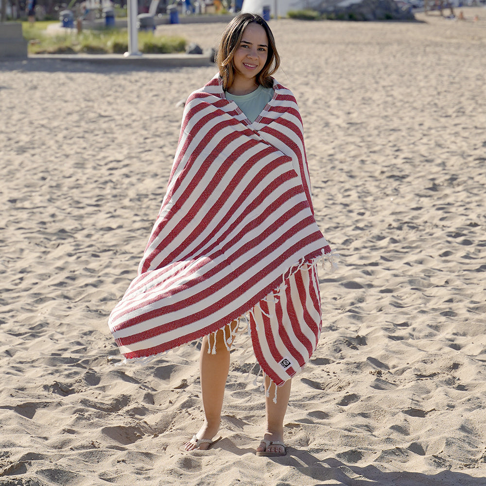 NIXY Acelia Bamboo Turkish Beach Towel, woman wrapped in a red and white striped bamboo-cotton towel on a sandy beach