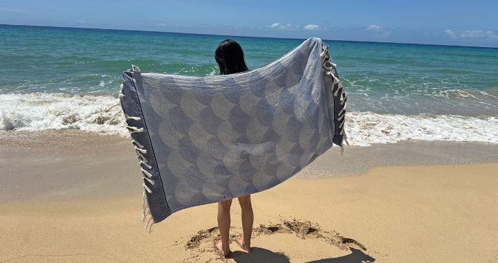 Women holding up NIXY Shell Turkish cotton beach towel in front of the water