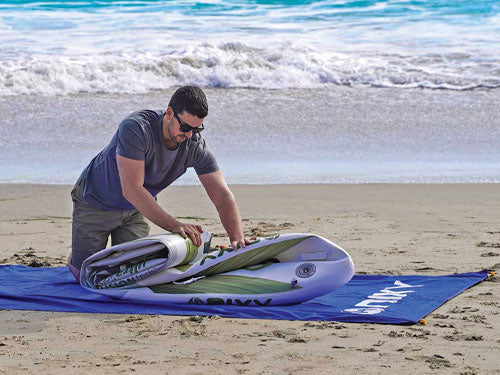 Man rolling up inflatable paddleboard on the sand at the beach with the water in the background.