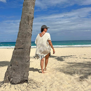 Person walking on a beach while wearing a women's swimsuit cover up with a palm tree and ocean view 