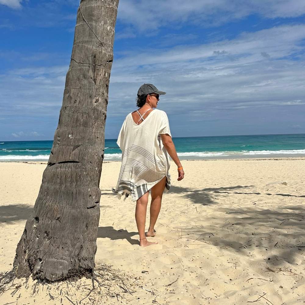 Person walking on a beach while wearing a women's swimsuit cover up with a palm tree and ocean view 