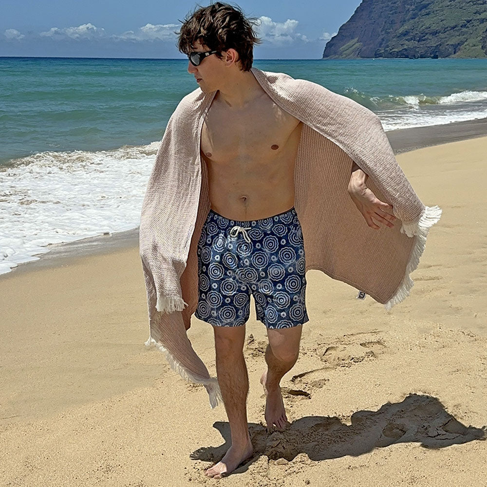 Man on a beach wearing blue patterned shorts and sunglasses, with a towel draped over his shoulder.