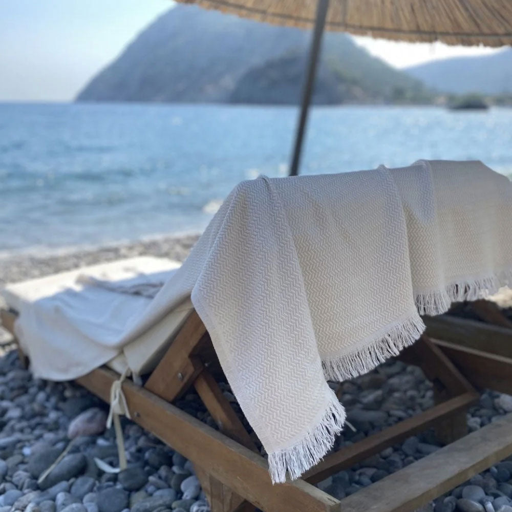 Beige towel draped over a wooden chair on a pebbly beach with ocean and mountains in the background