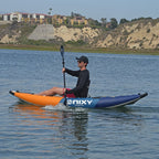 A man paddling an inflatable Nixy Tahoe 114 kayak on a calm lake, with sandy cliffs and residential buildings in the background.