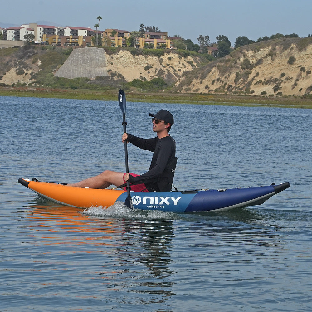 A man paddling an inflatable Nixy Tahoe 114 kayak on a calm lake, with sandy cliffs and residential buildings in the background.