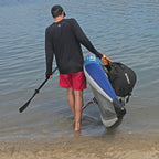 A man carries the lightweight NIXY Tahoe 114 Single inflatable kayak in shallow water on a sandy shore. He is wearing a UPF 50+ long-sleeve rash guard, red shorts, and a black cap, viewed from behind.