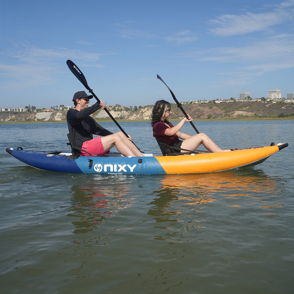 Two people paddle a NIXY tandem Tahoe 150 inflatable kayak on a calm body of water under a blue sky, with a shoreline and buildings visible in the background.