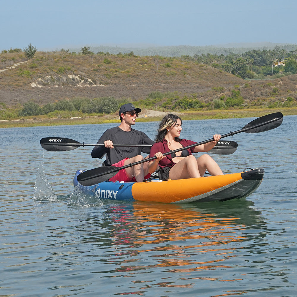 A couple paddling in a NIXY Tahoe 150 tandem inflatable kayak on calm water with a hilly, grassy shoreline in the background. The kayak is blue and orange with black paddle blades branded 'NIXY'.