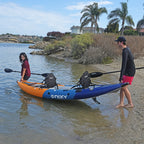 A couple pushes a Nixy Tahoe 150 two person inflatable kayak into shallow water on a sandy beach, with palm trees and houses in the background.