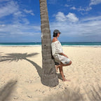 Person leaning against a palm tree wearing a cover up poncho on a sandy beach with ocean and sky in the background