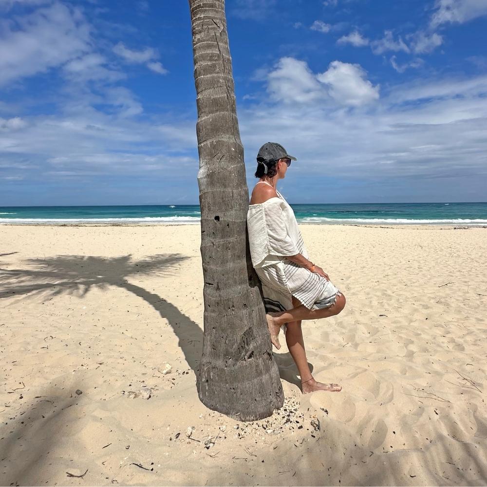 Person leaning against a palm tree wearing a cover up poncho on a sandy beach with ocean and sky in the background