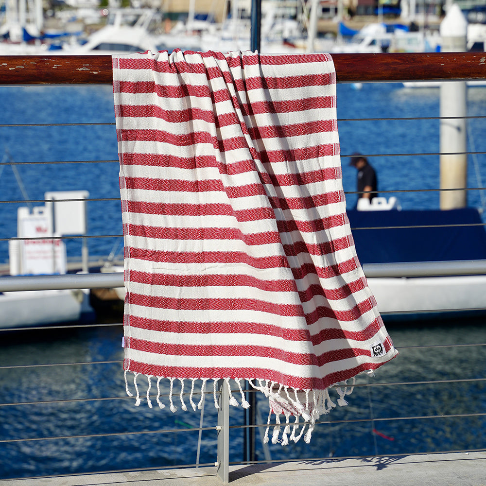 Red and white striped Turkish cotton beach towel with tassels hanging on a railing by a marina.