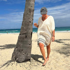 Woman in a white cover-up and cap standing on a beach with palm tree and ocean view.