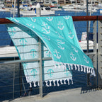 Turquoise Turkish cotton beach towel with white anchor pattern draped over a railing by a marina.