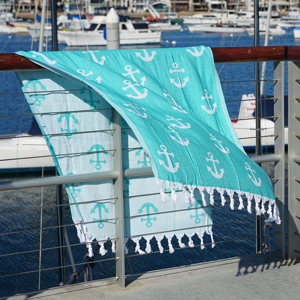 Turquoise Turkish cotton beach towel with white anchor pattern draped over a railing by a marina.