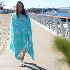 Woman wrapped in a turquoise Turkish cotton beach towel with anchor patterns on a beach boardwalk.