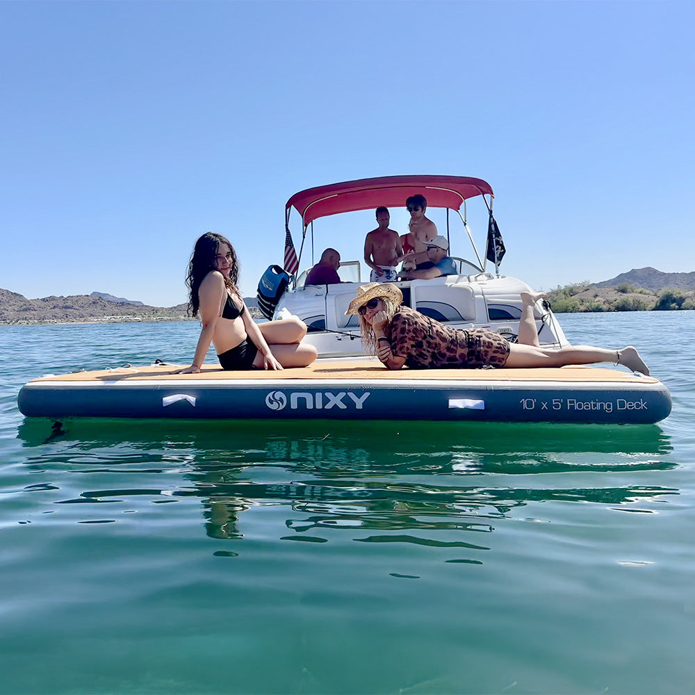 women sunbathing on nixy 5 by 10 foot inflatable floating deck in front of a boat on a sunny lake