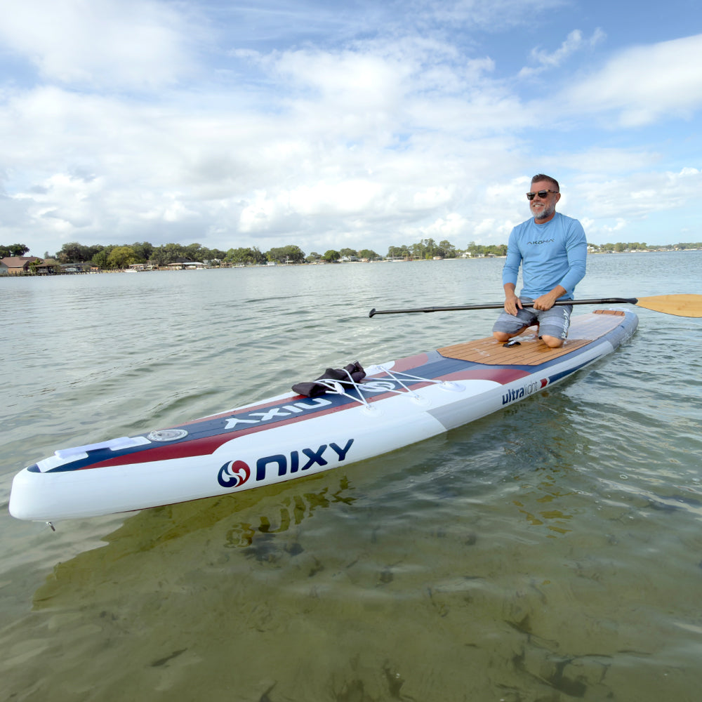 Man on a NIXY Malibu 14' Performance Inflatable Paddleboard on a calm body of water with a clear sky.
