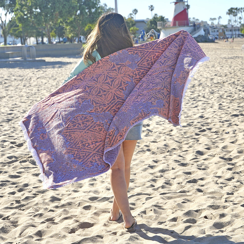 Person holding a patterned Turkish cotton beach towel on a sandy beach with a lighthouse in the background.