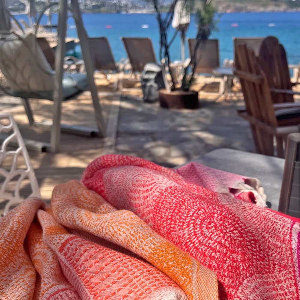 Close-up of a red and orange patterned towel with a blurred beach scene in the background.