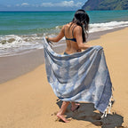 Woman on a beach holding a blue towel, with ocean and mountains in the background