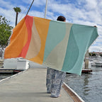 Person holding a NIXY Turkish beach towel by a dock with boats and palm trees in the background